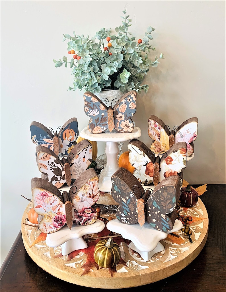May include: Six wooden butterfly figurines with autumn-themed floral patterns. The butterflies are arranged on a tiered wooden tray with white pedestals. The tray is decorated with autumn leaves and gourds.