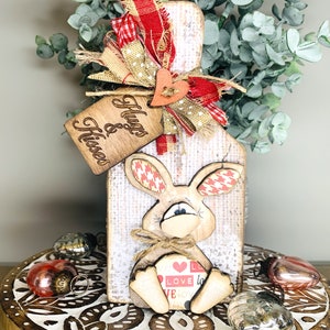 May include: Wooden bunny cutout with a red and white checkered pattern on its ears. The bunny is holding a heart with the word "love" on it. The bunny is attached to a wooden sign with the words "Hugs & Kisses" on it. The sign is decorated with a red and white checkered ribbon, a tan burlap ribbon, and a tan heart.