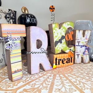 May include: Wooden Halloween-themed sign spelling out "Trick or Treat." The letters are decorated with patterned paper, ribbons, and Halloween motifs. A black pumpkin with "Spooky" written on it is in the background.