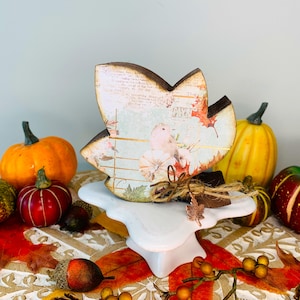 May include: A leaf-shaped wooden decoration with a bird and floral design, displayed on a white pedestal. The decoration has a brown border and is tied with twine. Pumpkins and fall foliage surround the decoration.