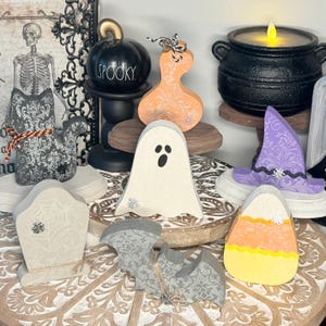 May include: A collection of wooden Halloween decorations including a black cat, a white ghost, a purple witch hat, a candy corn, a gray tombstone, a gray bat, and an orange gourd, arranged with a black cauldron and a black pumpkin labeled SPOOKY.