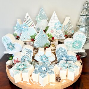 May include: A collection of decorative wooden Christmas ornaments in various shapes, including trees, hearts, and snowflake-like designs. The ornaments feature light blue and white patterns, with snowflake accents and small red berries. They are arranged on a wooden tray.