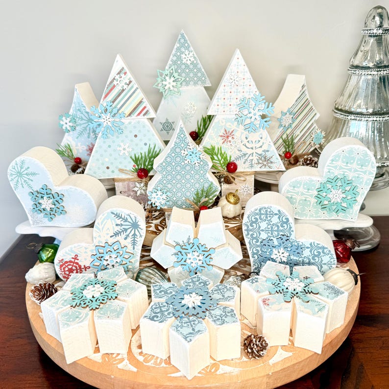 May include: A collection of decorative wooden Christmas ornaments in various shapes, including trees, hearts, and snowflake-like designs. The ornaments feature light blue and white patterns, with snowflake accents and small red berries. They are arranged on a wooden tray.