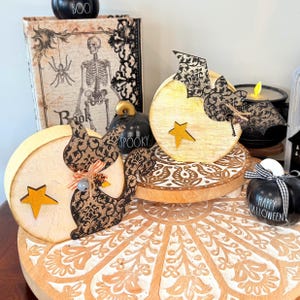 May include: Two round wooden decorations featuring a black cat and a bat with intricate black damask patterns. Each piece has a light wood finish, a small yellow star cutout, and decorative twine accents. These items are displayed on a tiered tray.