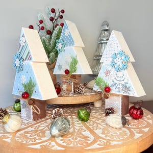 May include: Wooden Christmas tree decorations with light blue snowflake patterned tops and wooden bases. Each tree is adorned with a wooden snowflake, faux greenery, and red berries. Decorative ornaments and pinecones are arranged around the trees.