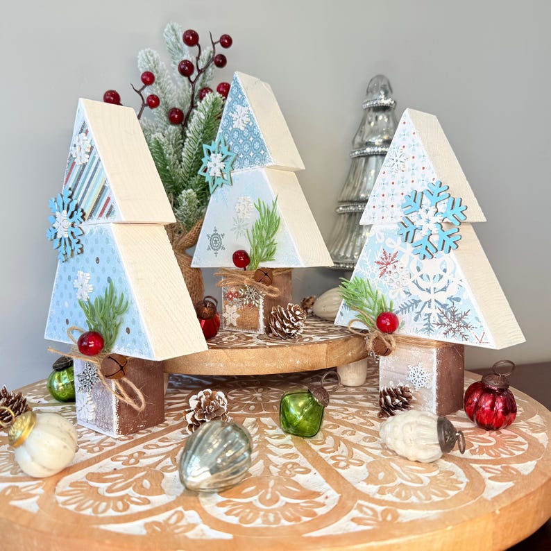 May include: Wooden Christmas tree decorations with light blue snowflake patterned tops and wooden bases. Each tree is adorned with a wooden snowflake, faux greenery, and red berries. Decorative ornaments and pinecones are arranged around the trees.