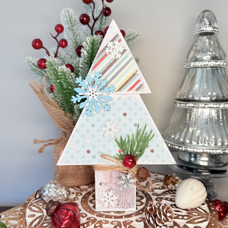 May include: A wooden Christmas tree decoration with a light blue background and snowflake accents. The tree has a striped top section and is adorned with faux greenery, red berries, and a small bell. The base is a small wooden block.