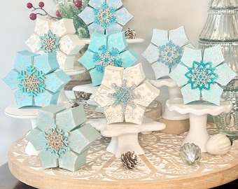 May include: A collection of seven dimensional snowflake ornaments in shades of light blue, white, and teal. Each snowflake features a central decorative embellishment with glittery accents and intricate patterns, displayed on small white pedestals.