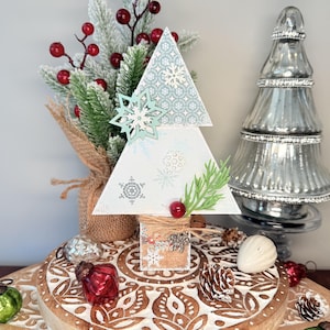 May include: A decorative wooden Christmas tree with a snowflake design. The tree is white with light blue snowflake accents and a light blue snowflake ornament. It is decorated with faux greenery, red berries, and a small bell. The tree sits on a wooden surface.