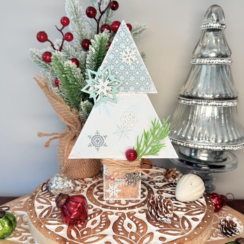 May include: A decorative wooden Christmas tree with a snowflake design. The tree is white with light blue snowflake accents and a light blue snowflake ornament. It is decorated with faux greenery, red berries, and a small bell. The tree sits on a wooden surface.