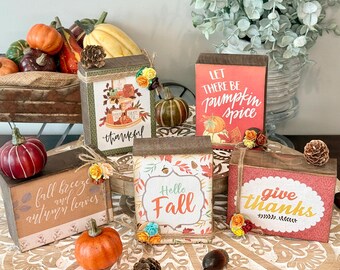 May include: Four rectangular wooden blocks with autumn-themed designs and text including 'thankful', 'let there be pumpkin spice', 'fall breeze and autumn leaves', 'hello fall', and 'give thanks', decorated with small artificial flowers and pinecones.