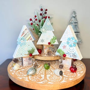 May include: Wooden Christmas tree decorations with snowflake and patterned paper designs. The trees are decorated with faux greenery, red berries, and wooden snowflake accents. The trees are displayed on a wooden tray with decorative ornaments and pinecones.