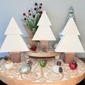 May include: Three wooden Christmas tree decorations with a light cream finish, each with a square base wrapped in twine. The trees are displayed on a decorative wooden tray with Christmas ornaments and pinecones.