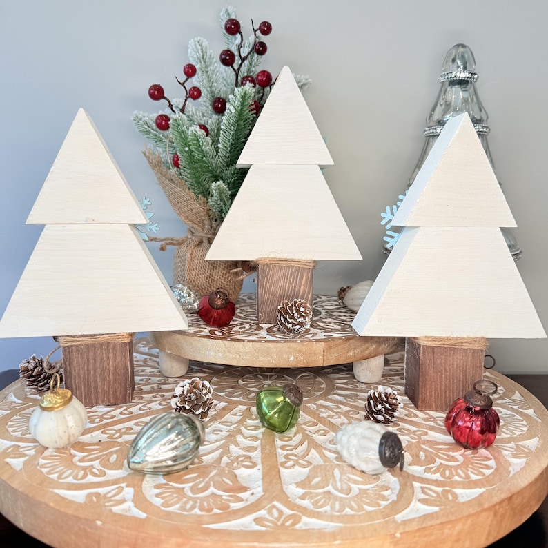 May include: Three wooden Christmas tree decorations with a light cream finish, each with a square base wrapped in twine. The trees are displayed on a decorative wooden tray with Christmas ornaments and pinecones.