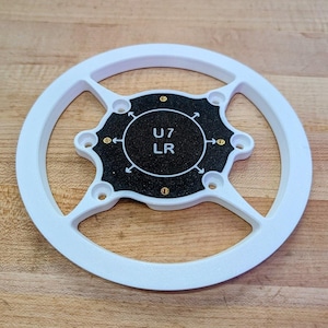 May include: A white circular plastic frame with a central black textured plate featuring the text U7 LR and four directional arrows. The assembly includes four small brass-colored metal inserts positioned around the central black circular section.