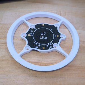 May include: A white circular mounting bracket with a black center plate featuring the text U7 Lite, 1G JB, 4" R JB, and 3" R JB. The bracket includes four brass-colored threaded inserts and multiple screw holes for installation purposes.