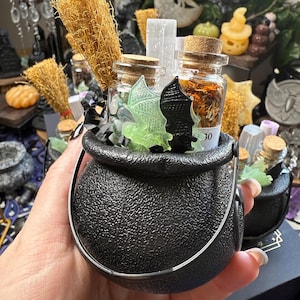 May include: A black cauldron with a textured surface, filled with small glass bottles, dried herbs, and decorative elements. Miniature brooms and bat-shaped accents are included. The number "30" is visible on one of the bottles. The cauldron is held in a hand.