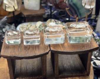 May include: Four clear, square glass candle holders of varying sizes arranged in a row. Each holder features a circular indentation on the top surface. The glass holders are positioned on two dark brown wooden stands with open, rectangular bases.
