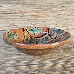 May include: A handmade, shallow ceramic bowl with a wide rim. The bowl features a brown base with a painted design of teal leaves and brown branches. The rim is a darker brown color, and the bowl has a rustic, aged appearance.