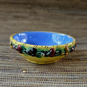 May include: A small, ceramic bowl with a bright blue interior and a yellow exterior. The rim is decorated with a green band featuring red and brown fruit. The bowl has a textured, woven pattern on the outside.