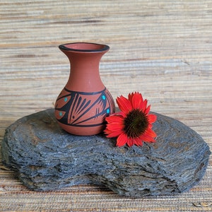 May include: A small, terracotta vase with a black rim and decorative black and turquoise geometric patterns. A vibrant red flower with a dark center rests beside the vase on a gray stone. The vase is approximately 4 inches tall.