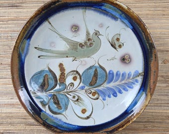 Könnte beinhalten: Runder Keramikteller mit handgemaltem Design. Der Teller zeigt einen Vogel und einen Schmetterling mit blauen und braunen floralen Elementen. Der Rand hat einen dunkelblauen und braunen Rand. Der Teller wird auf einem Holzständer präsentiert.