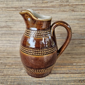 May include: A brown ceramic pitcher with a handle and spout. The pitcher has a glossy finish and is decorated with horizontal bands of small, raised dots. The interior of the spout is a light beige color. The pitcher is likely used for serving beverages.