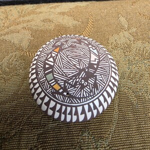 May include: A round, flat ceramic disc featuring a dark brown and white geometric pattern with fine lines. The design includes stylized lizard figures and small accents in muted orange, blue, and yellow, with a border of repeating white shapes.