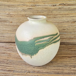 May include: A round ceramic vase with a speckled, off-white base and a green and tan abstract design. The vase has a wide, rounded body and a narrow opening at the top. The background is a textured, woven surface.