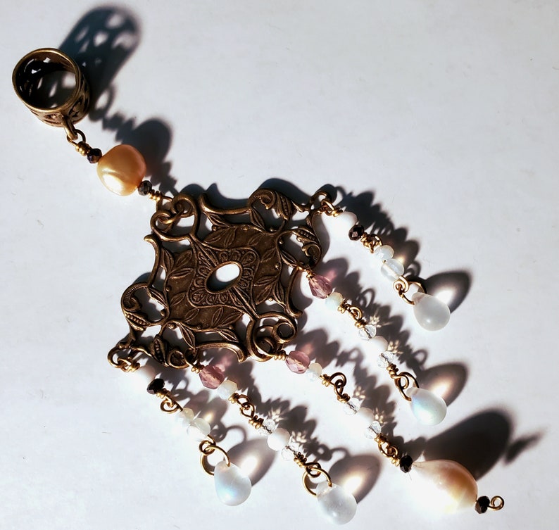 May include: A gold-toned pendant with a filigree design and multiple strands of beads. The beads are white, pink, and iridescent.