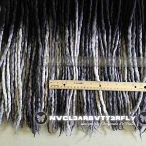 May include: A collection of ombre dreadlocks transitioning from black at the top to light gray at the ends. A wooden ruler is placed horizontally across the strands, showing a measurement of 12 inches. Text reads NVCL3ARBVTT3RFLY designs by Cheyenne Le Hale.