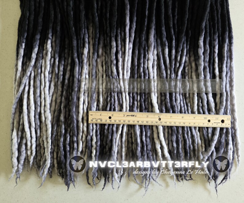 May include: A collection of ombre dreadlocks transitioning from black at the top to light gray at the ends. A wooden ruler is placed horizontally across the strands, showing a measurement of 12 inches. Text reads NVCL3ARBVTT3RFLY designs by Cheyenne Le Hale.