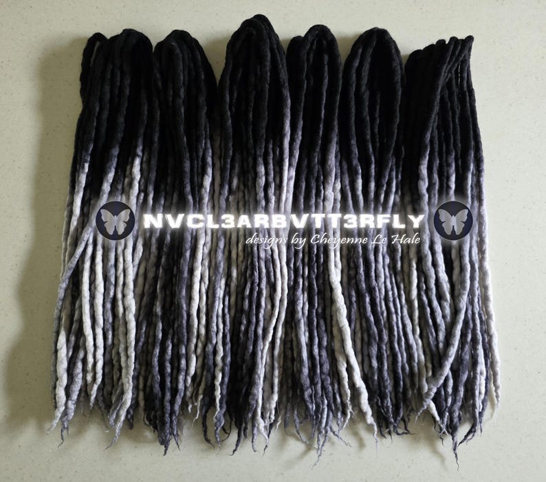 May include: A set of synthetic dreadlocks featuring a black to grey ombre color gradient. The hair extensions are arranged in parallel rows. Overlaid text reads NVCL3ARBVTT3RFLY designs by Cheyenne Le Hale with two butterfly icons on the sides.