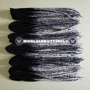 May include: A set of synthetic dreadlocks featuring an ombre color transition from solid black at the roots to a mottled grey and white at the tips. The text NVCL3ARBVTT3RFLY designs by Cheyenne Le Hale is centered over the dreadlocks.