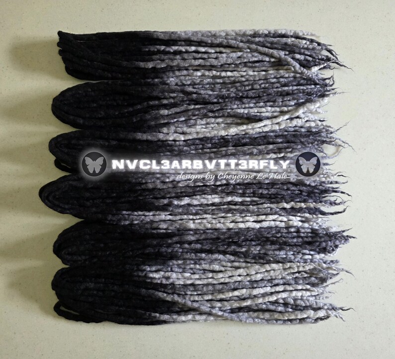 May include: A set of synthetic dreadlocks featuring an ombre color transition from solid black at the roots to a mottled grey and white at the tips. The text NVCL3ARBVTT3RFLY designs by Cheyenne Le Hale is centered over the dreadlocks.