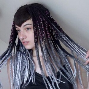 May include: A set of synthetic dreadlocks featuring a gradient color scheme that transitions from deep black at the roots to shades of charcoal gray and light silver at the tips. The individual strands are textured with a braided or wrapped pattern.
