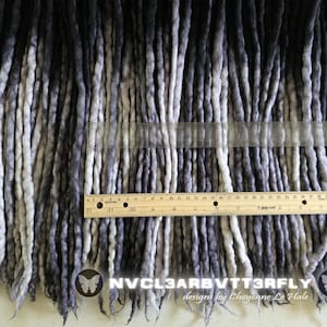 May include: A collection of ombre synthetic dreadlocks transitioning from black to grey and white. A wooden ruler is placed horizontally across the center, showing a measurement of 12 inches. The text NVCL3ARB VTT3RFLY designs by Cheyenne Le Hale is visible.