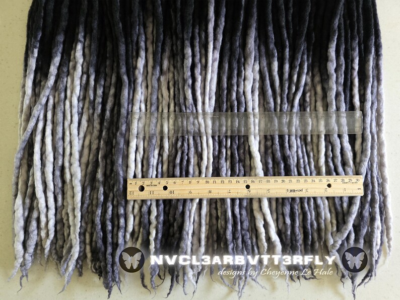 May include: A collection of ombre synthetic dreadlocks transitioning from black to grey and white. A wooden ruler is placed horizontally across the center, showing a measurement of 12 inches. The text NVCL3ARB VTT3RFLY designs by Cheyenne Le Hale is visible.