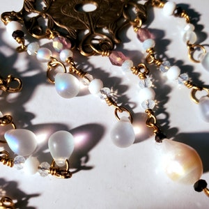 May include: A gold-toned metal pendant with a floral design and dangling iridescent white teardrop-shaped beads. The pendant is adorned with small white and pink beads.