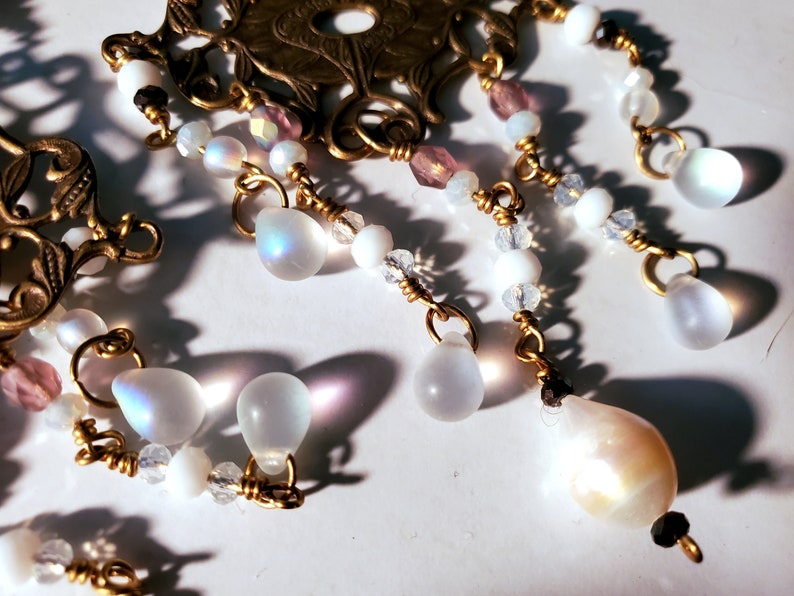 May include: A gold-toned metal pendant with a floral design and dangling iridescent white teardrop-shaped beads. The pendant is adorned with small white and pink beads.