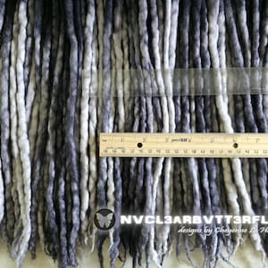 May include: A collection of synthetic dreadlocks in a gradient of black, charcoal, and white. A wooden ruler is placed horizontally across the strands, showing a width of 9 inches. The text NVCL3ARBVTT3RFLY designs by Cheyenne Le Hale is visible.