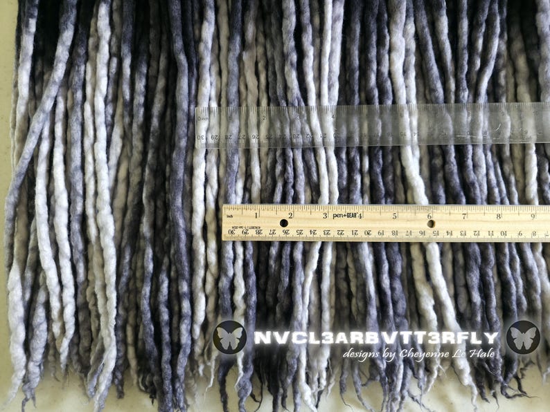 May include: A collection of synthetic dreadlocks in a gradient of black, charcoal, and white. A wooden ruler is placed horizontally across the strands, showing a width of 9 inches. The text NVCL3ARBVTT3RFLY designs by Cheyenne Le Hale is visible.