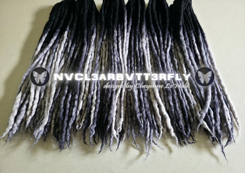 May include: A collection of synthetic dreadlocks featuring an ombre color transition from black at the roots to a light grey at the tips. The text NVCL3ARBVTT3RFLY designs by Cheyenne Le Hale is centered across the strands with butterfly icons.