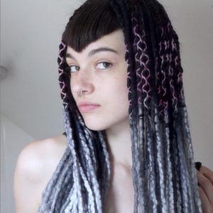 May include: A set of long hair extensions featuring black roots with pink and white interwoven thread detailing, transitioning into textured grey and white yarn-like strands. The extensions are styled with straight dark bangs and a septum ring.