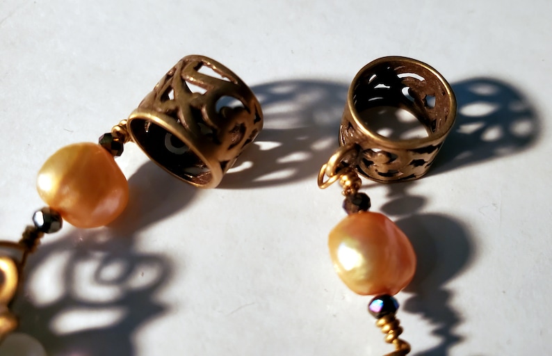 May include: A gold necklace with two ornate metal spacers and two pearlescent beads. The spacers have a filigree design.