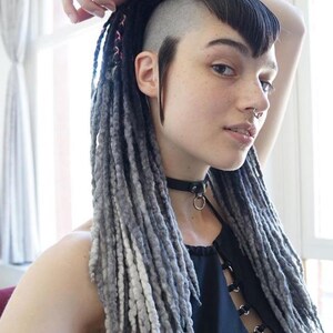 May include: A black choker necklace with a central metal ring, worn with a black sleeveless top featuring a front lace-up design with silver eyelets. The hair is styled in long, textured gray and black dreadlocks with a shaved side and bangs.