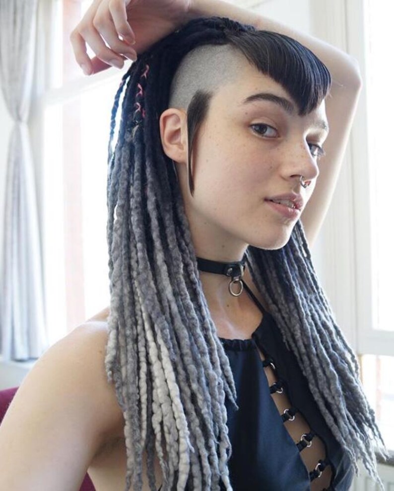 May include: A black choker necklace with a central metal ring, worn with a black sleeveless top featuring a front lace-up design with silver eyelets. The hair is styled in long, textured gray and black dreadlocks with a shaved side and bangs.