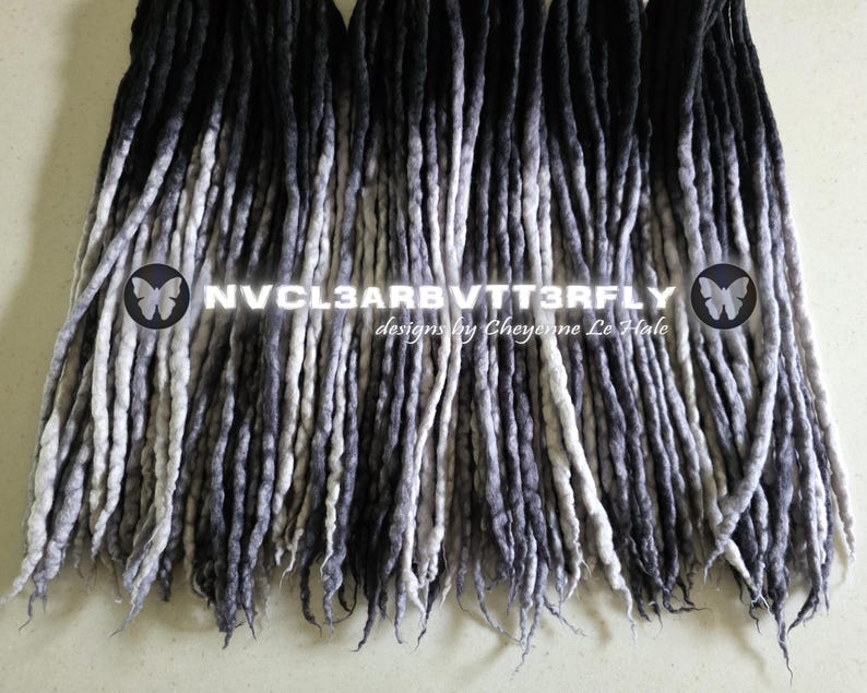 May include: A collection of synthetic dreadlocks featuring a gradient color transition from black at the roots to light gray at the tips. The text NVCL3ARBVTT3RFLY and designs by Cheyenne Le Hale are overlaid on the center of the dreadlocks.