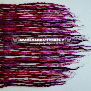 Op de afbeelding: Een verzameling handgemaakte dreadlocks in tinten rood, paars en roze, gerangschikt op een wit oppervlak. De dreadlocks zijn van verschillende lengtes en diktes, met een getextureerd uiterlijk. De afbeelding bevat de tekst "NVCL3ARBVTT3RFLY designs by Cheyenne Le Hale".