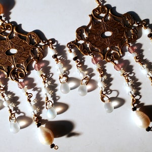May include: A pair of gold-tone earrings with a delicate, ornate design. Each earring features a central pendant with a floral motif and a series of dangling beads, including white, pink, and iridescent beads.